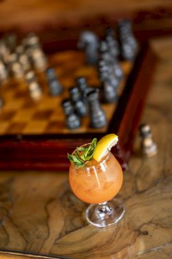 A glass cocktail with orange drink, mint and a lemon wedge, with blurred bar bottles in the background.