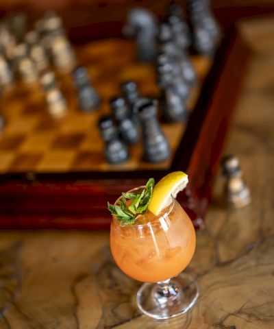 A glass cocktail with orange drink, mint and a lemon wedge, with blurred bar bottles in the background.