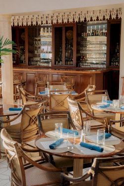 Cozy restaurant with wooden bar, wicker chairs, round tables set with blue napkins and water glasses, a decorative shelf in the background.