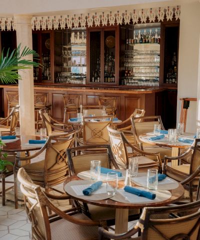 Cozy restaurant with wooden bar, wicker chairs, round tables set with blue napkins and water glasses, a decorative shelf in the background.