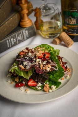 A fresh salad on a white plate: greens, strawberries, feta or goat cheese, nuts, and crumbled topping; wine and corked bottle in the background.