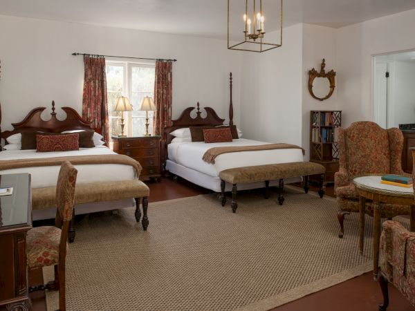 Two large four-poster beds with matching wooden nightstands, a seating area with upholstered chairs, a small desk, and a cozy rug in a vintage hotel room.