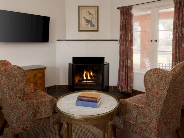 A cozy living room with a fireplace, two patterned armchairs, a round coffee table, a wall-mounted TV, and sliding glass doors opening to a patio.
