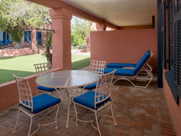 A covered patio with a round table and white chairs with blue cushions, plus a blue lounge chair by the wall.