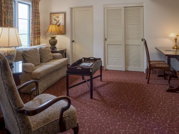 A cozy hotel room with a sofa, armchairs, a coffee table, a desk, lamps, a window, and closed double doors in a warm, traditional decor.