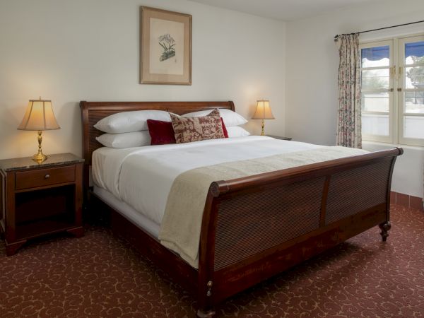 A neatly made hotel-style bedroom with a wooden bed, two nightstands with lamps, a framed art piece, and patterned carpet near a window with curtains.