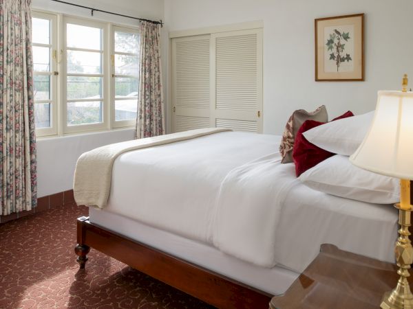 A tidy hotel-style bedroom with a large wooden bed, white bedding, plaid curtains, a bedside lamp, and a small dresser by the window.