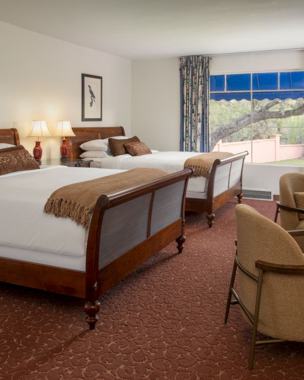 A hotel room with two large beds, a seating area, desk, lamps, and a window with blue curtains overlooking a sunny view.