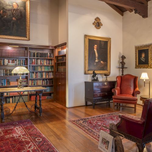 A cozy library and sitting room with tall bookshelves, red upholstered chairs, wooden desk, lamps, and framed portraits on the walls, warm and inviting.