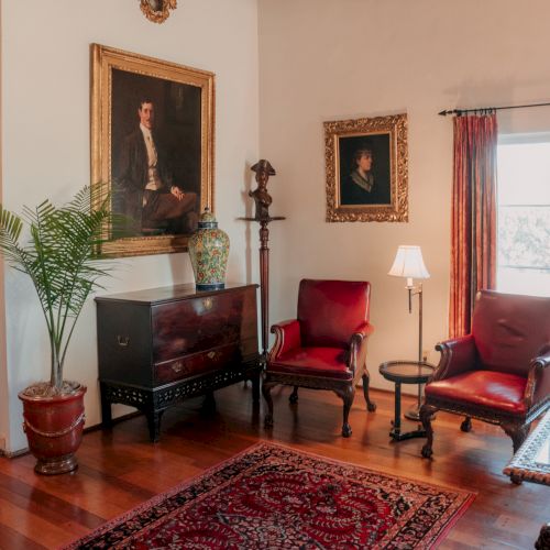 A classic parlor with antique portraits, a wooden cabinet, red upholstered chairs, a Persian rug, potted plant, and warm lighting.