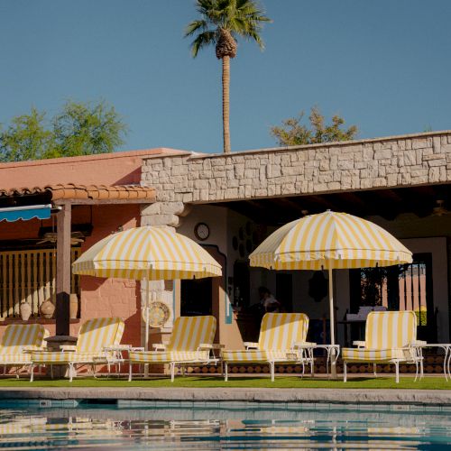 A resort-style pool scene with yellow lounge chairs under striped umbrellas, a stone building, and a tall palm tree against a clear blue sky.