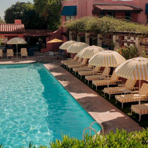 A resort pool scene with a bright blue swimming pool, beige striped lounge chairs, and yellow umbrellas, surrounded by greenery and pink buildings.