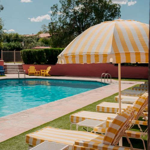 A sunny pool scene with yellow-striped umbrellas and lounge chairs by a bright blue pool, a grassy yard, and a red-pink wall in the background.