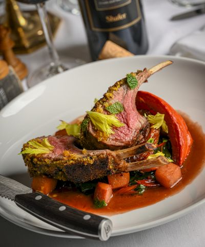 A plated rack of lamb with herbs, roasted vegetables, and a rich tomato-based sauce in a fine dining setting.