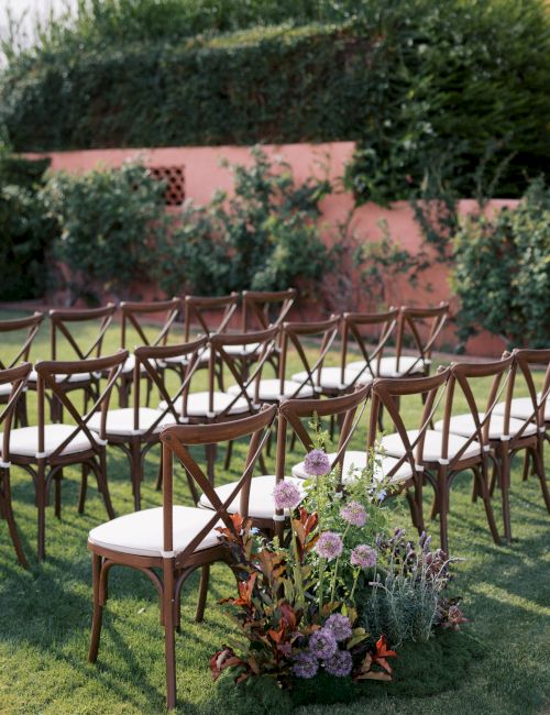 An outdoor ceremony setup with rows of wooden chairs, white cushions, and a garden flower arrangement on the grass.