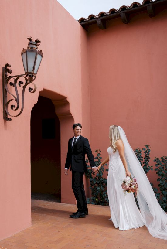 A bride and groom holding hands outside a peach stucco building, the bride in a white gown with bouquet in hand.