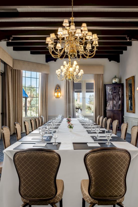 An elegant dining room with a long set table, gilded chandeliers, tall curtains, and ornate decor.