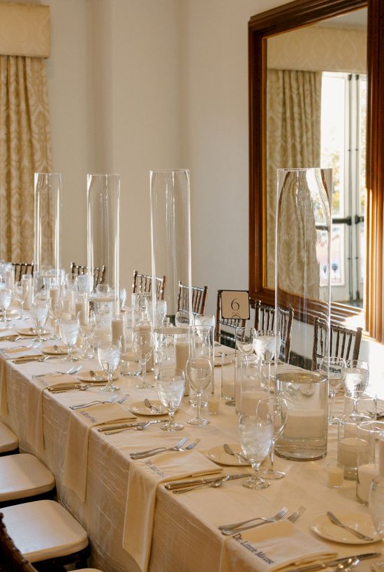 A long banquet table set for a formal meal with white tablecloths, numerous wine glasses, silverware, and folded napkins, in a bright room with windows.