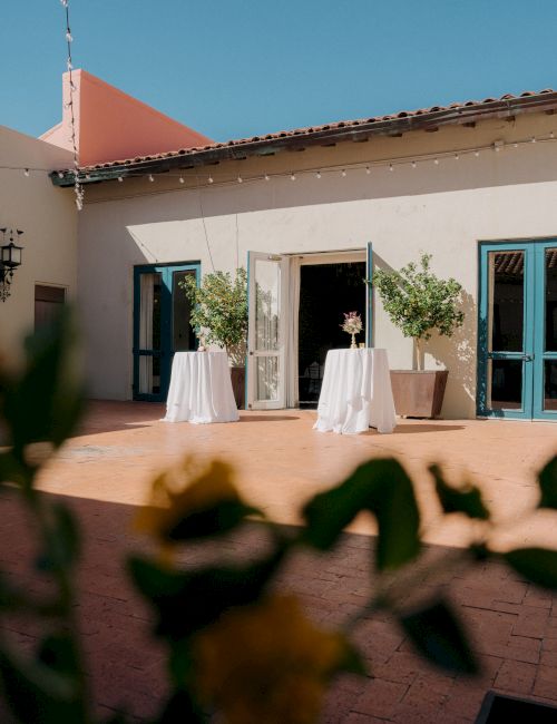Two small tables with white tablecloths and potted trees sit on a sunny patio in front of a light-colored building with blue doors.