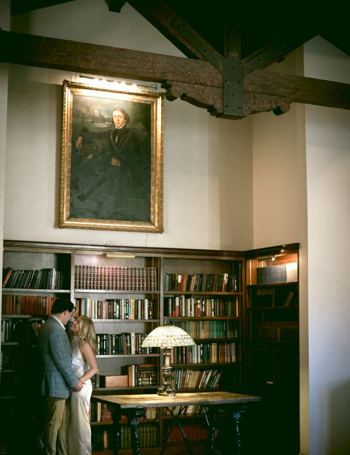 Couple stands by a bookshelf in a cozy library, candle-lit table, portrait above, warm wooden beams, vintage atmosphere.