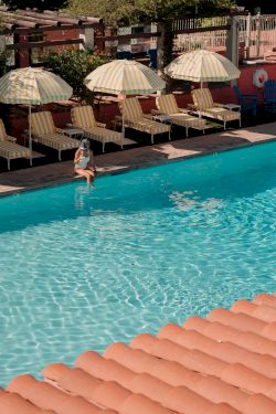 A sunny pool scene with clear blue water, sun loungers under striped umbrellas, and a person stepping into the pool, end it.