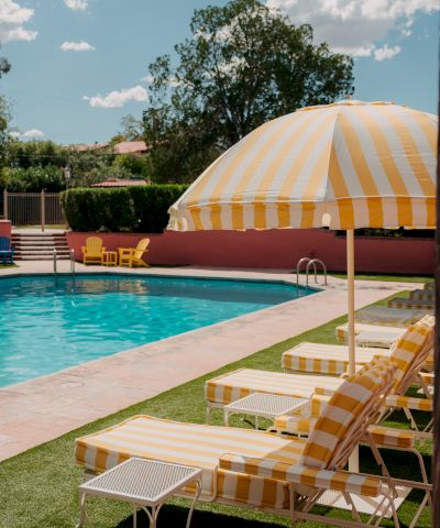 A sunny poolside scene with striped yellow and white lounge chairs, a matching umbrella, and a bright blue pool under a clear sky.