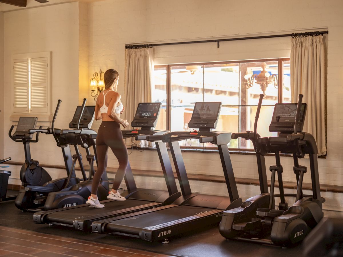 A woman works out on treadmills in a sunny gym, framed by windows and beige curtains.