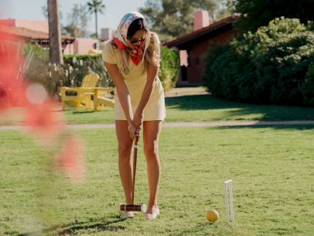 A woman in a light yellow top and white shorts tees off on a sunny golf course, focused on her swing with a ball and tee set ahead.