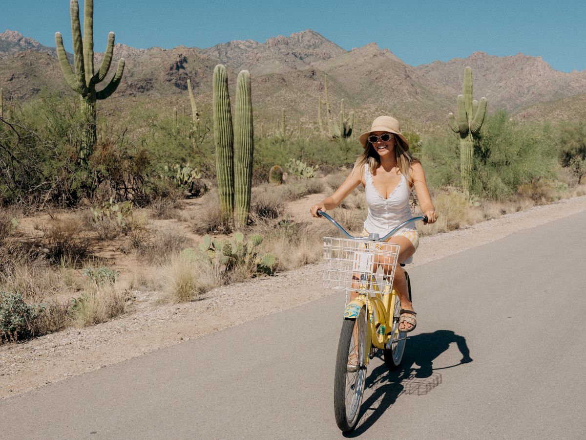 A woman rides a small yellow bicycle on a desert road, wearing a hat and white outfit, with tall cacti and rugged mountains in the background.