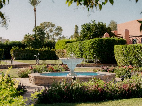 A lush garden with a central stone fountain, trimmed hedges, flowering plants, and a sunny courtyard vibe under leafy trees. ending now.