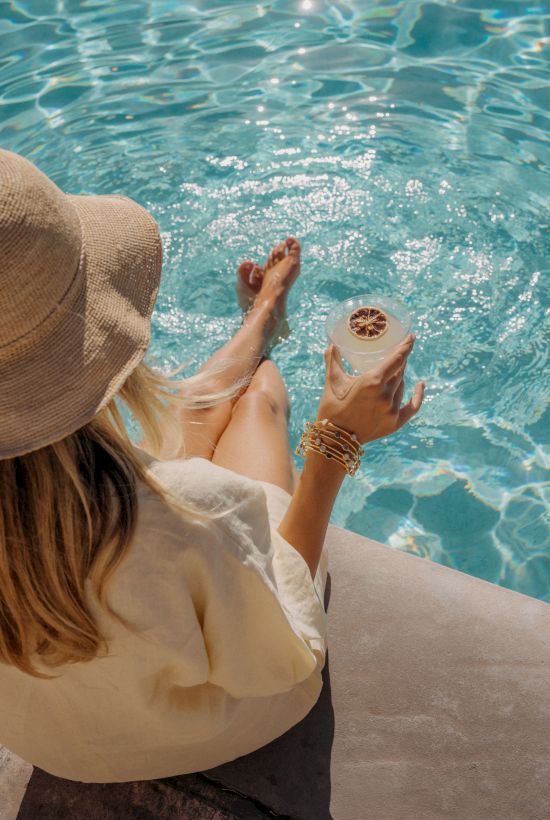 A woman wearing a sunhat sits by a bright pool, holding a tropical drink and dipping her feet into the clear blue water to relax.