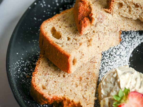 Sliced rustic bread on a dark plate with a dollop of cream and a strawberry garnish, light dusting of powdered sugar, coffee cup in background.