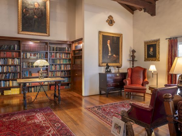 A cozy library and lounge: bookshelves, a wooden desk with lamp, classic paintings, red upholstered chairs, and warm lamps on wood floors.