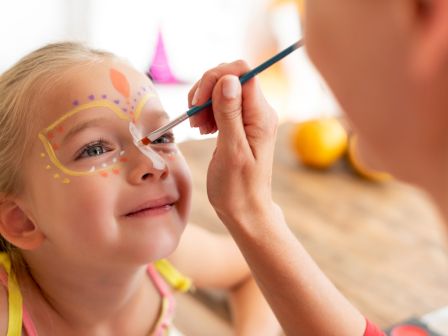A young girl has face paint being applied around her eyes by a smiling artist, colorful dotted designs around her cheeks and forehead.
