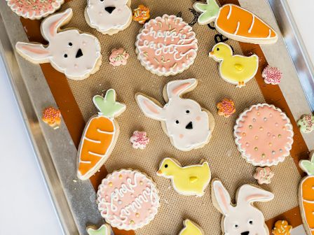 A baking sheet with Easter-themed cookies: white bunnies, carrot shapes, pink round cookies, plus small chick cookies and leaves.