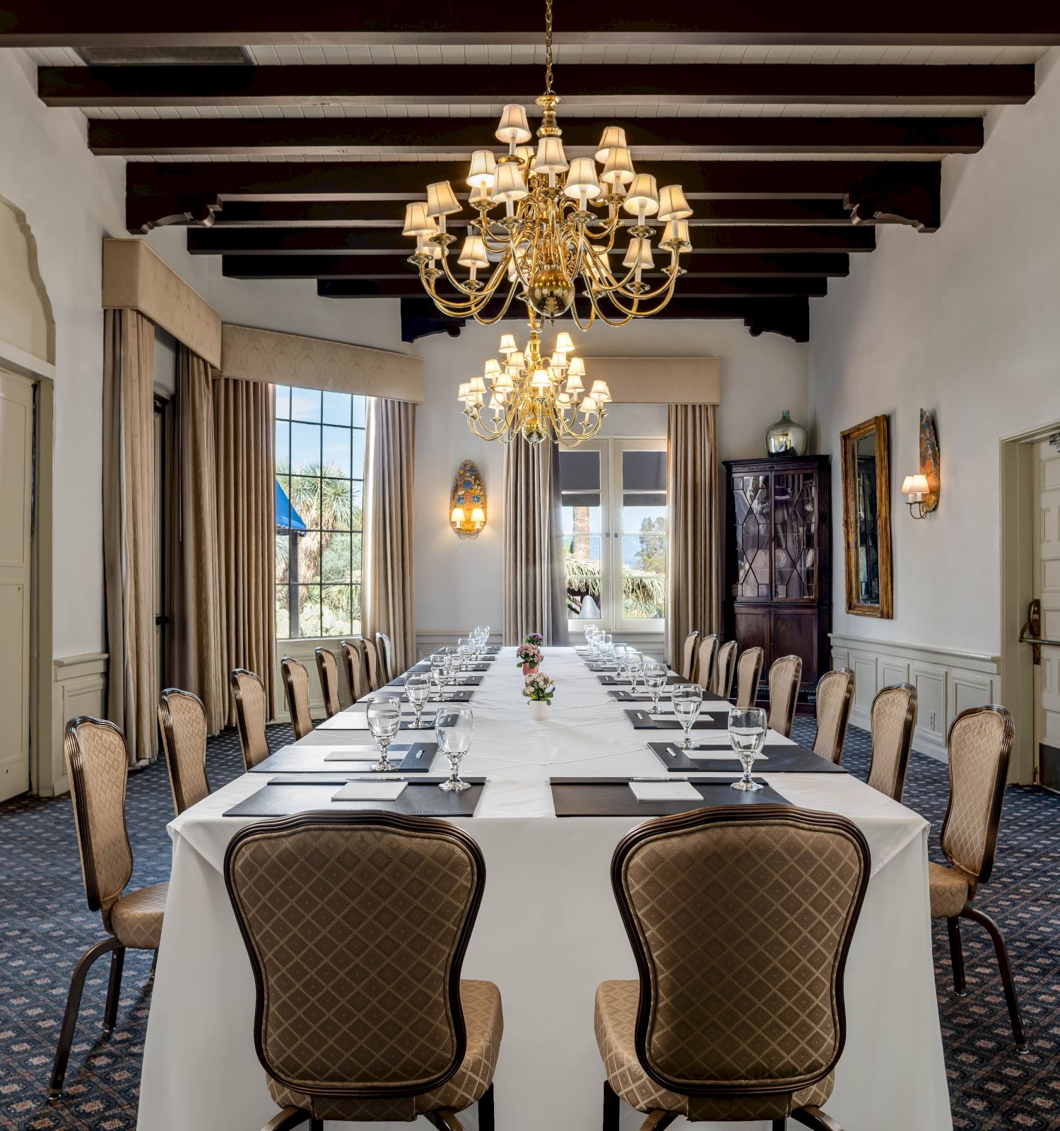 An elegant banquet room set with a long table, chairs, and grand gold chandeliers above.