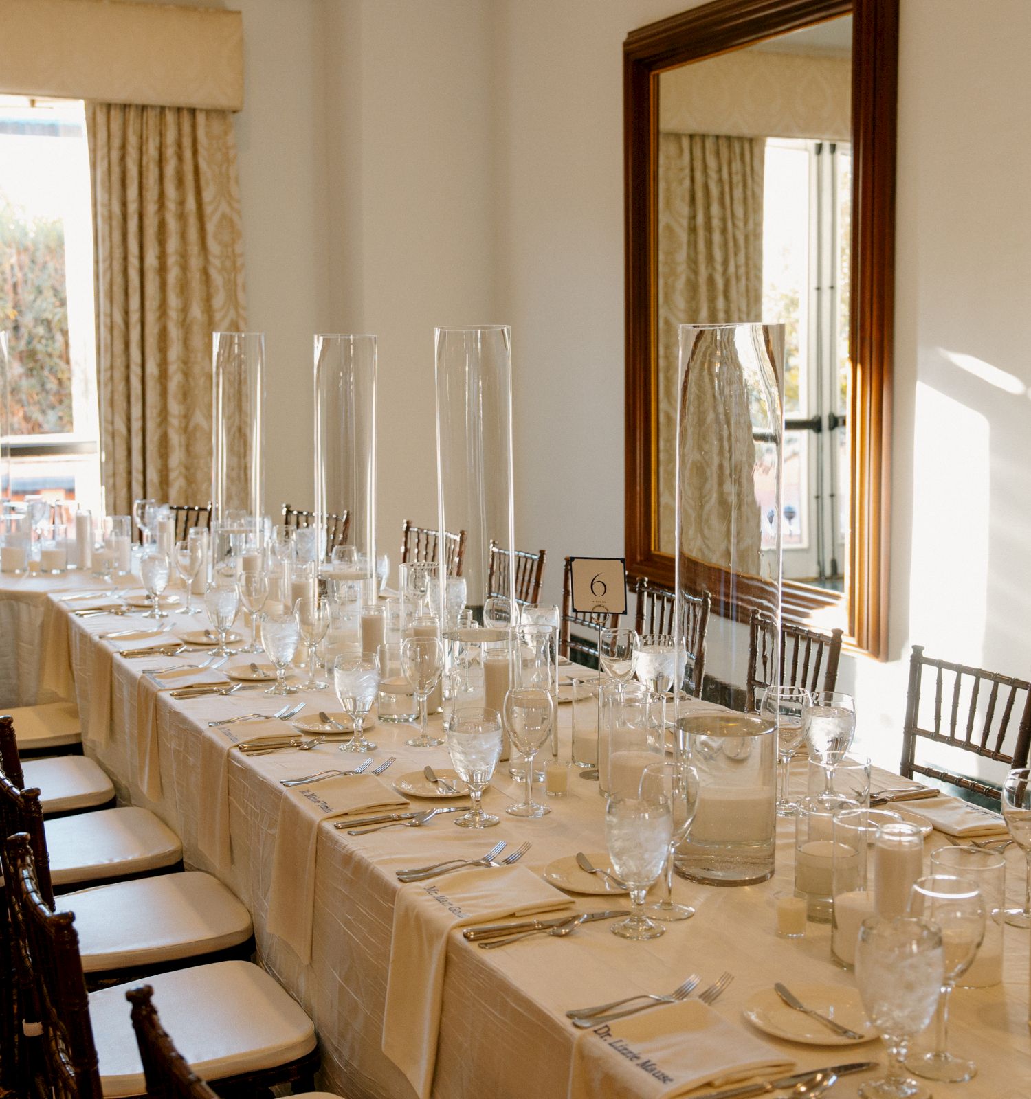 A long dining table set for a formal meal with white linens, numerous glasses, plates, and cutlery, in a bright room.