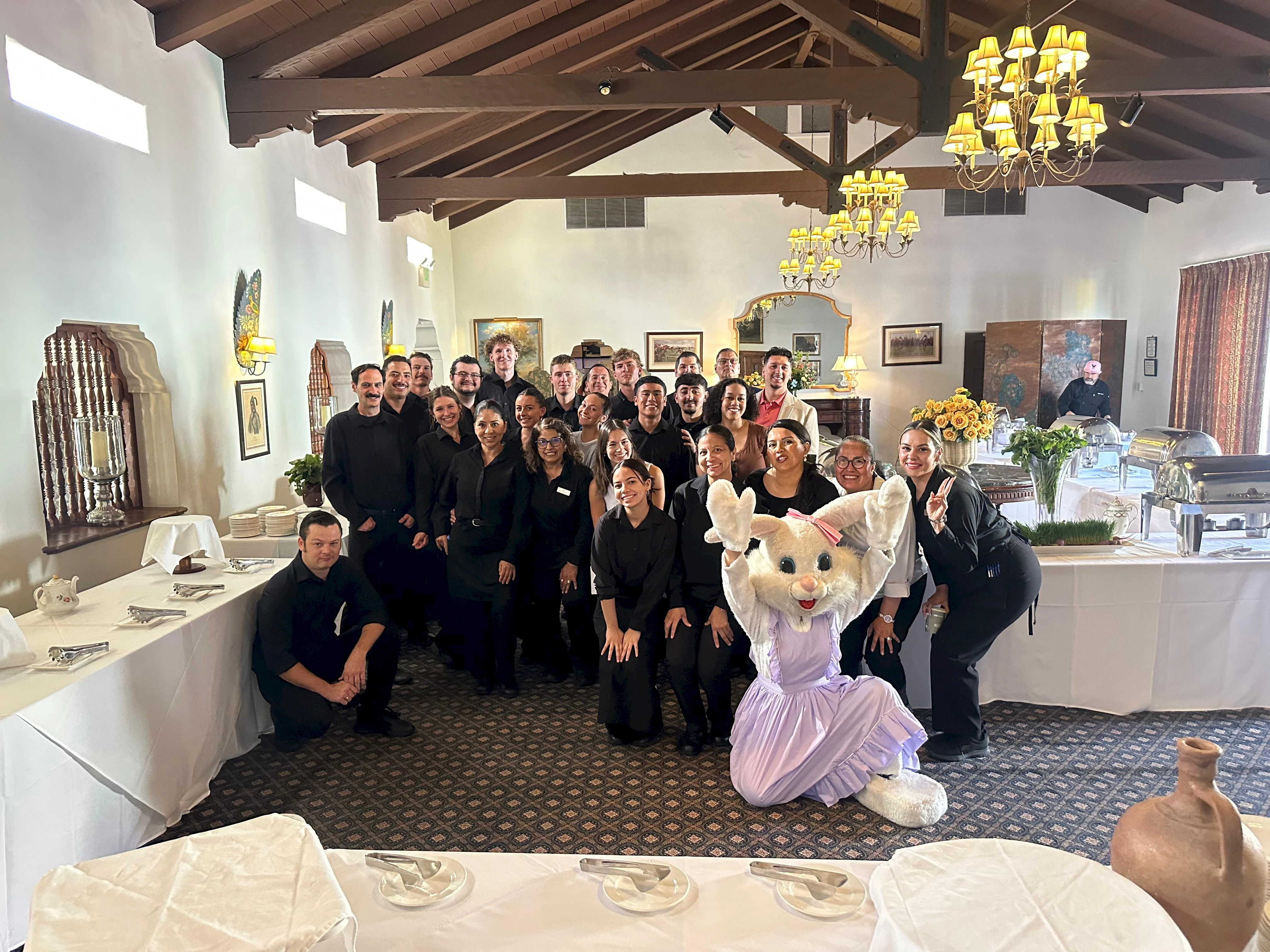 A large group photo with guests in a banquet hall, a smiling mascot in a purple dress posing center, tables set for a event.
