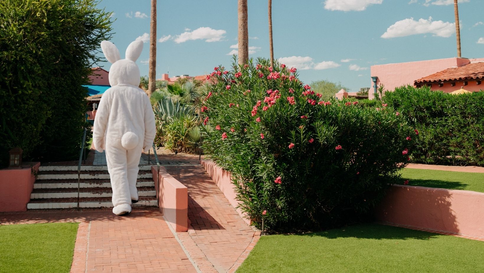 A person dressed in a white bunny costume walking along a brick path past flowering bushes and neatly trimmed lawns under a sunny sky.