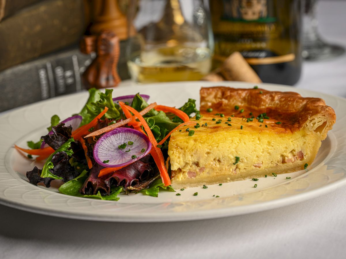 A plated slice of savory quiche with a side salad of mixed greens, radish, and carrots, garnished with herbs on a white plate.
