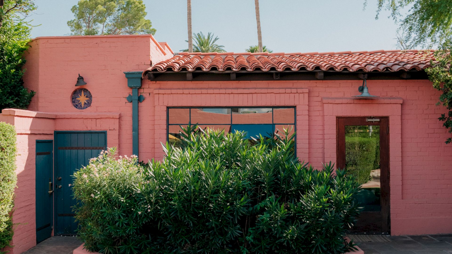 A pink stucco building with a red-tiled roof, blue doors, lush green shrubs in front, and a sunny, desert-like vibe.