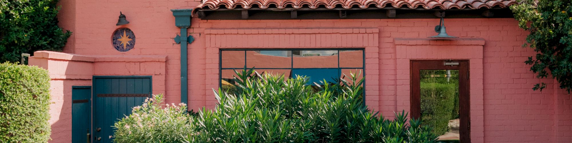 A pink stucco building with a red-tiled roof, blue doors, lush green shrubs in front, and a sunny, desert-like vibe.