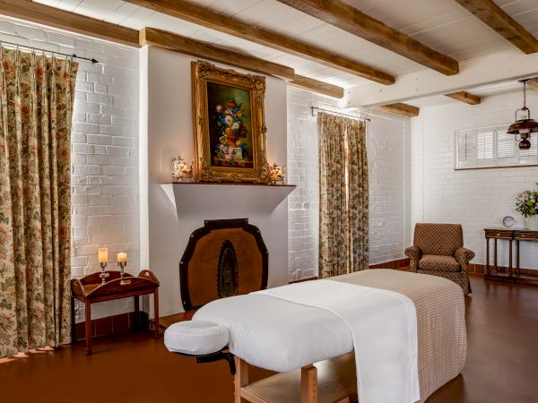 A cozy, vintage-style bedroom with a bed, floral curtains, framed art, candleholders, and wooden beams.
