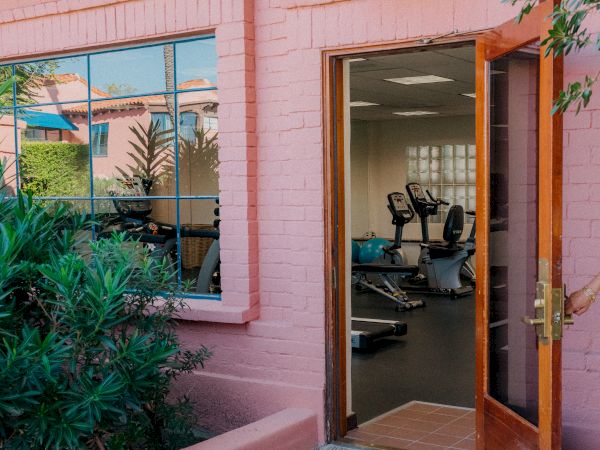 Outdoor view of a pink building with a wooden door open to a gym, visible exercise equipment inside, tropical plants nearby, sunny day.
