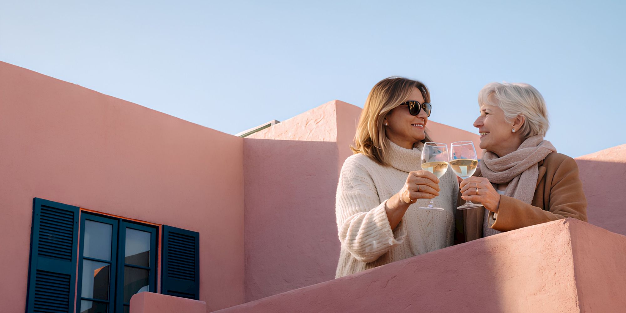 Two women on a pink balcony toast with wine glasses, enjoying a sunny outdoor moment together.