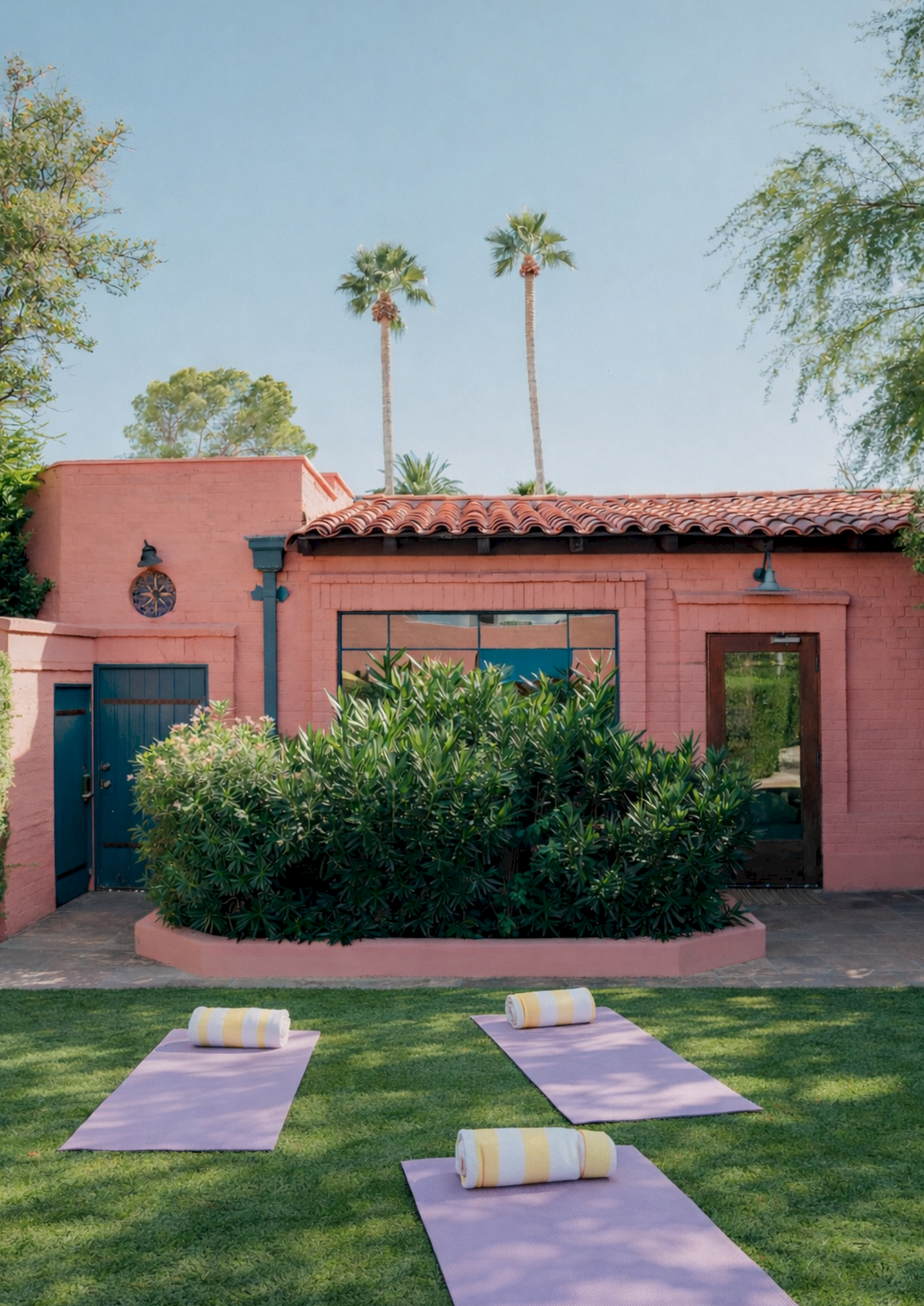 Outdoor retreat at a pink stucco villa with glass doors, lush greenery, and neatly laid lavender yoga mats with towels. End.