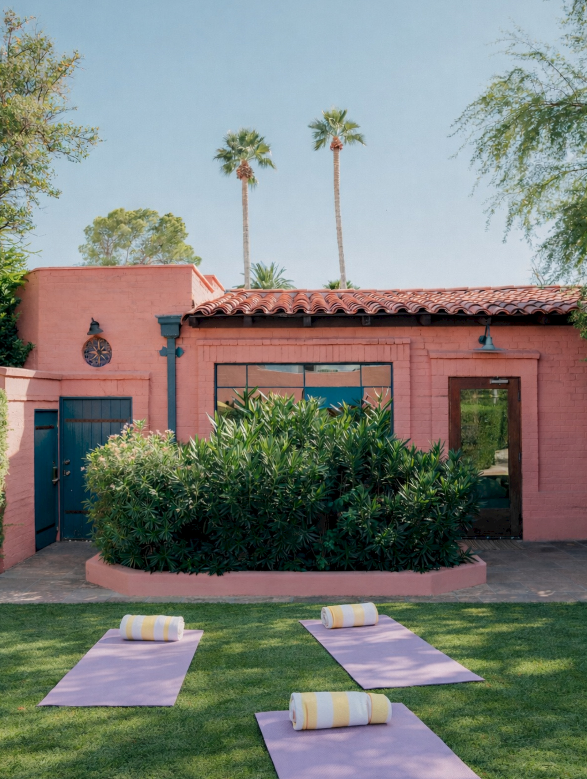 Outdoor retreat at a pink stucco villa with glass doors, lush greenery, and neatly laid lavender yoga mats with towels. End.