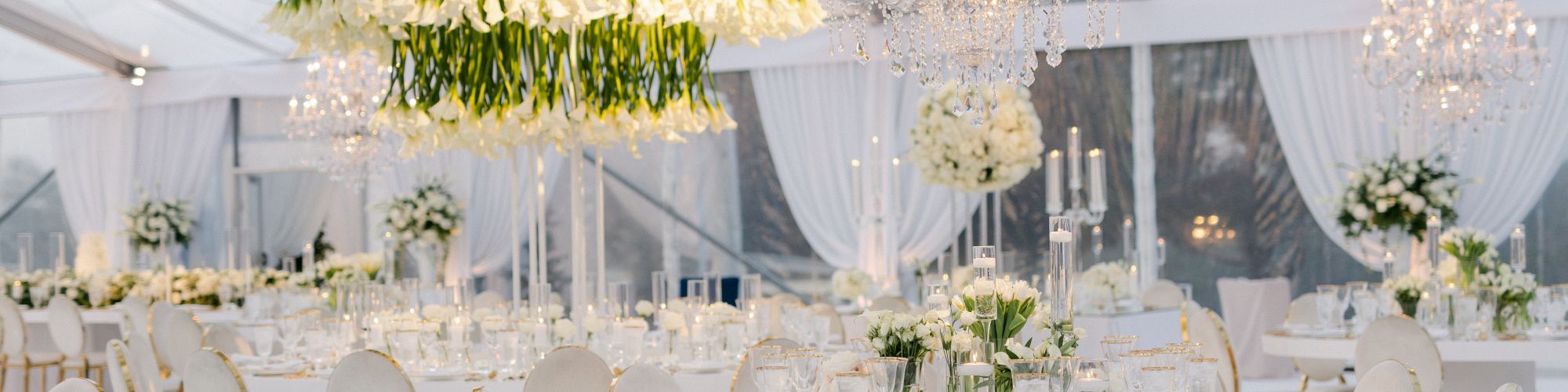 A luxurious wedding reception setup in a bright white tent with elegant gold-trimmed chairs, lush floral centerpieces, and chandeliers, dreamy and pristine.