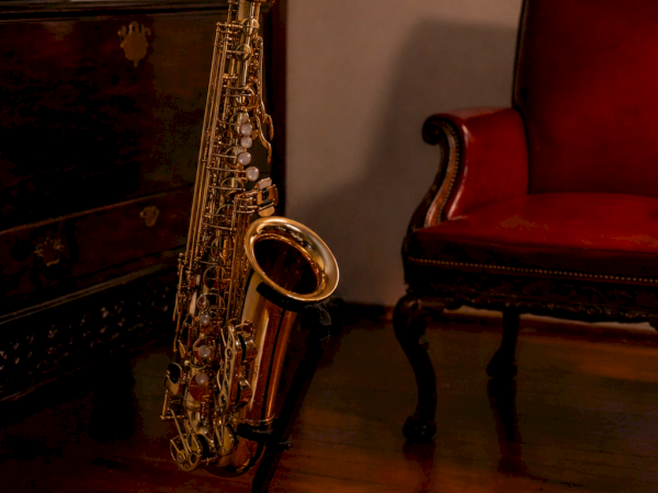 A gold saxophone on a stand in a dim, vintage room with a red chair, ornate rug, and antique furniture in the background.
