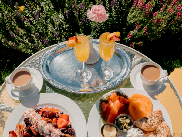 A sunny outdoor breakfast with two plates: pancakes and berries on one, assorted spreads and bread on the other; two orange cocktails and coffee cups on a silver tray, all set on a glass table.